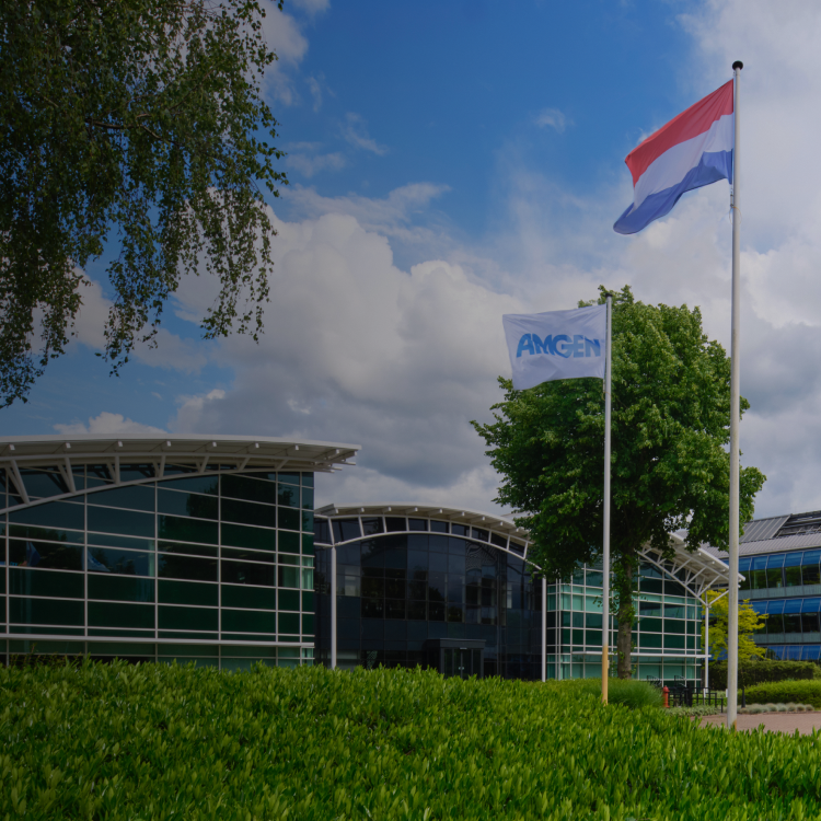Voorgevel van een modern Amgen-kantoorgebouw met glazen gevels, een Amgen-vlag wapperend aan een vlaggenmast, en het woord 'Support' in beeld. De lucht is deels bewolkt en de omgeving is groen en verzorgd.