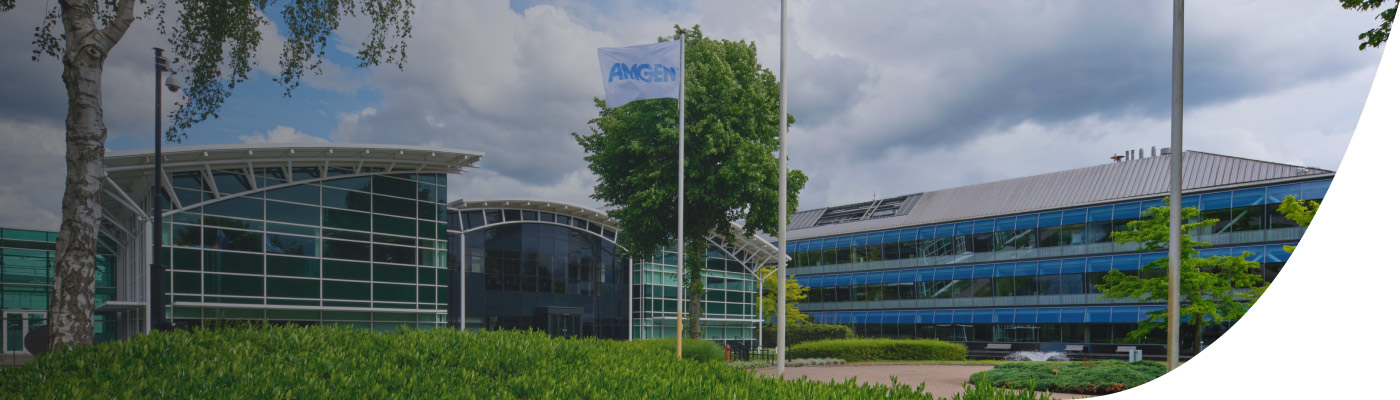 Voorgevel van een modern Amgen-kantoorgebouw met glazen gevels, een Amgen-vlag wapperend aan een vlaggenmast, en het woord 'Support' in beeld. De lucht is deels bewolkt en de omgeving is groen en verzorgd.